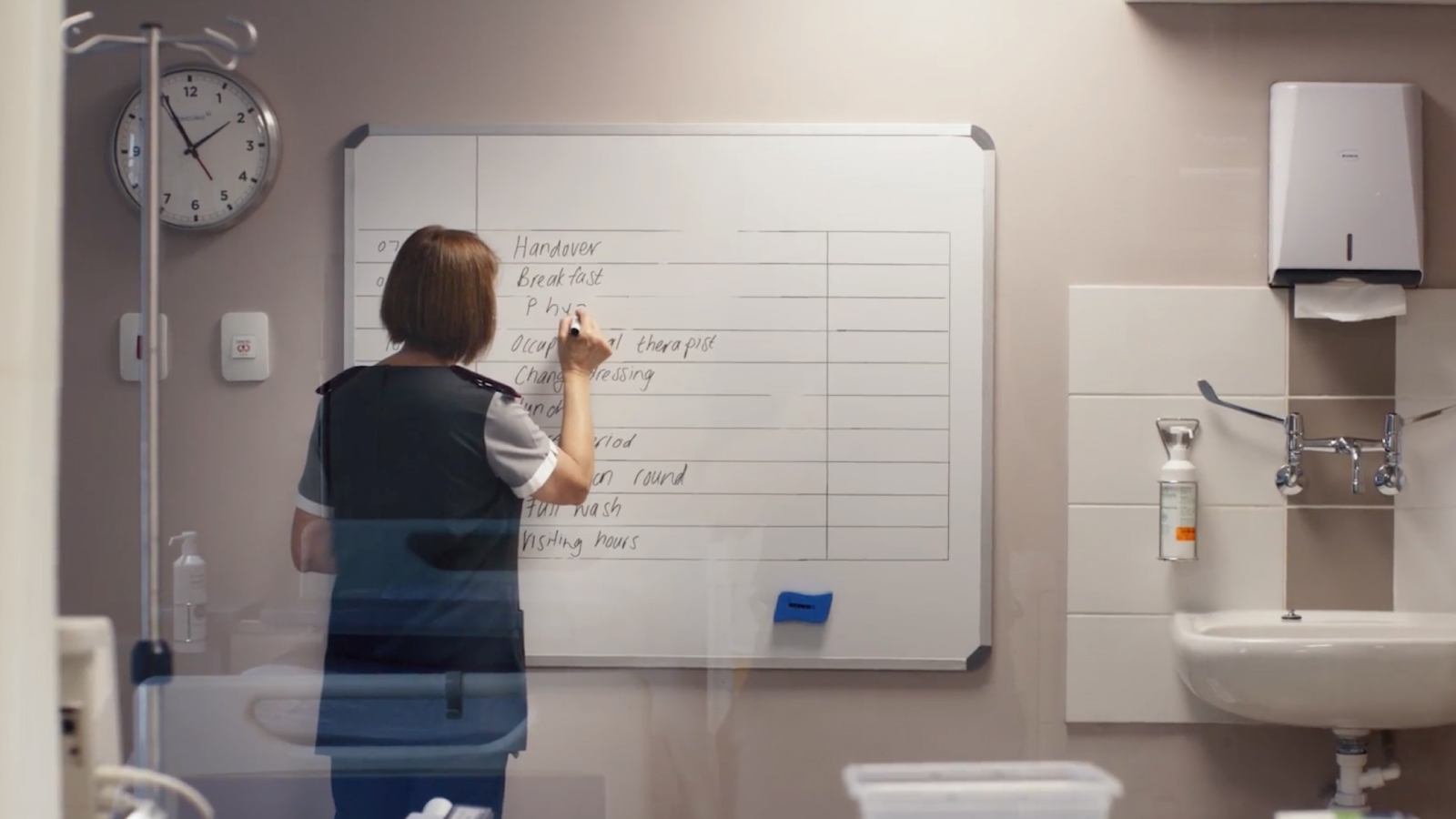 A healthcare professional writing on a whiteboard in a hospital setting, with a clock and medical equipment visible in the background.