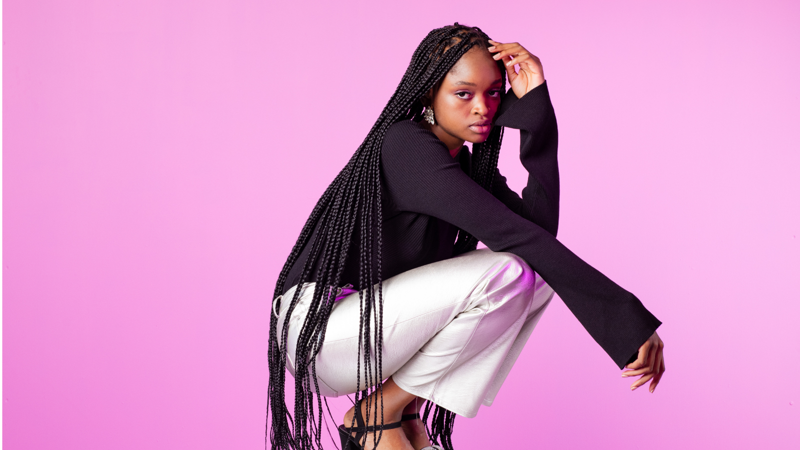 A woman with long braided hair crouching in a confident pose against a vibrant pink background, wearing a black top and metallic silver pants.