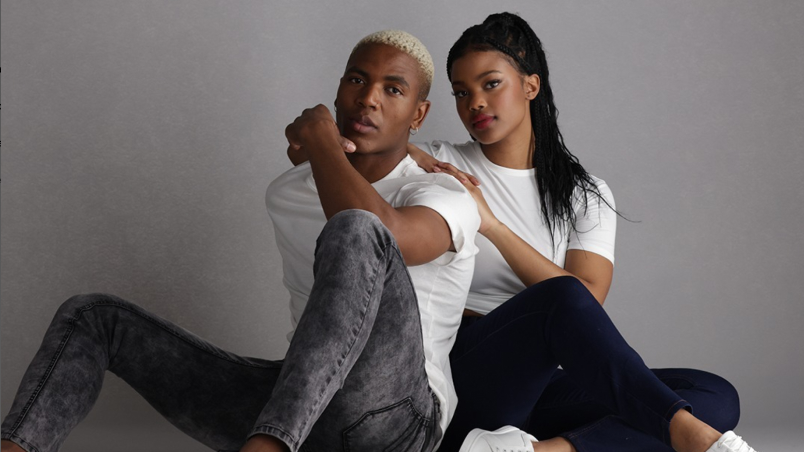 A stylish young man and woman sitting against a neutral gray background, both wearing matching white t-shirts and dark jeans, exuding a modern, casual aesthetic.