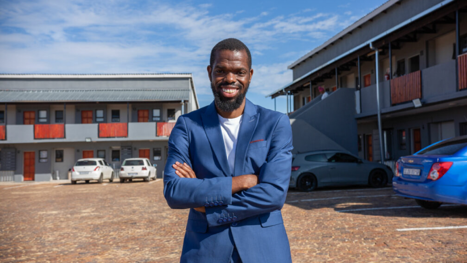 A man in a blue suit smiling with his arms crossed in front of a housing complex.