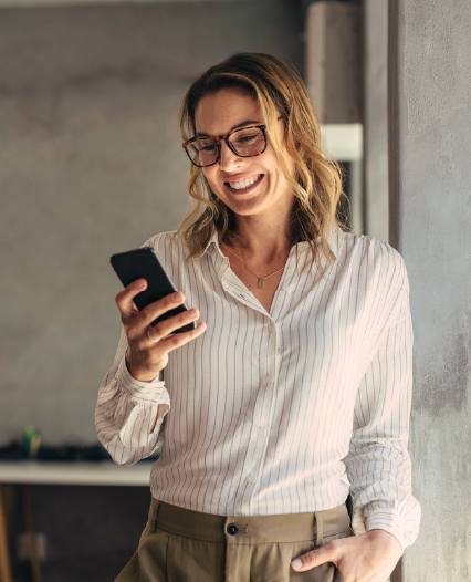 A smiling woman wearing glasses holds her phone, radiating joy and engagement in a bright setting.