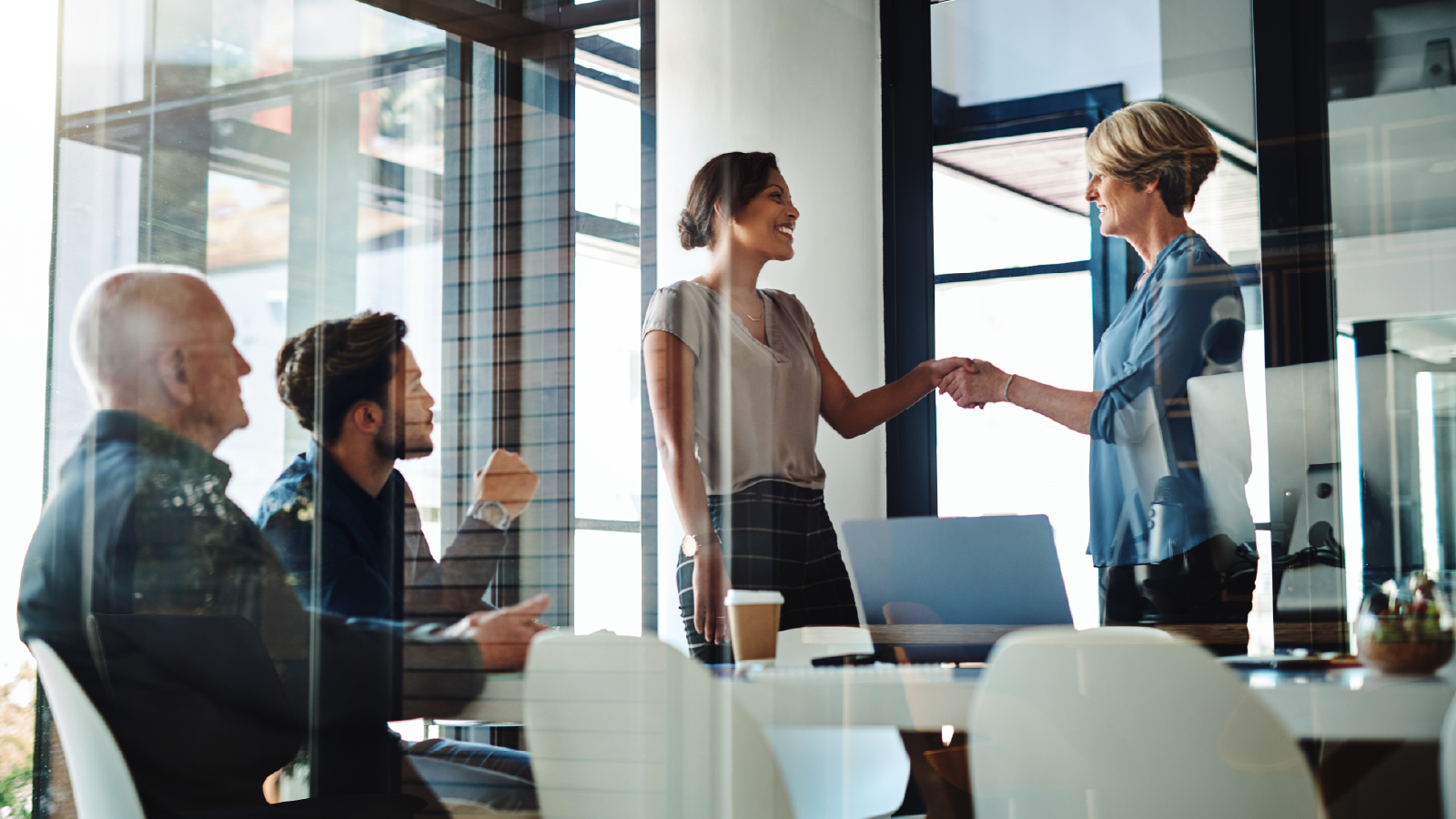 A business meeting in a glass-walled office, with two women shaking hands while colleagues observe.