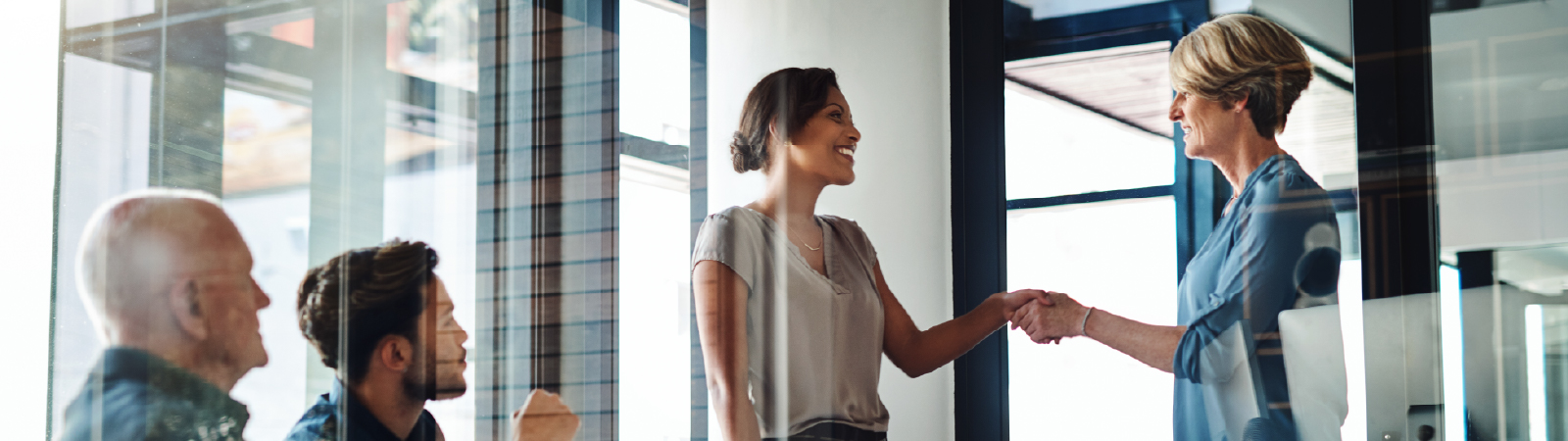 A business meeting in a glass-walled office, with two women shaking hands while colleagues observe.