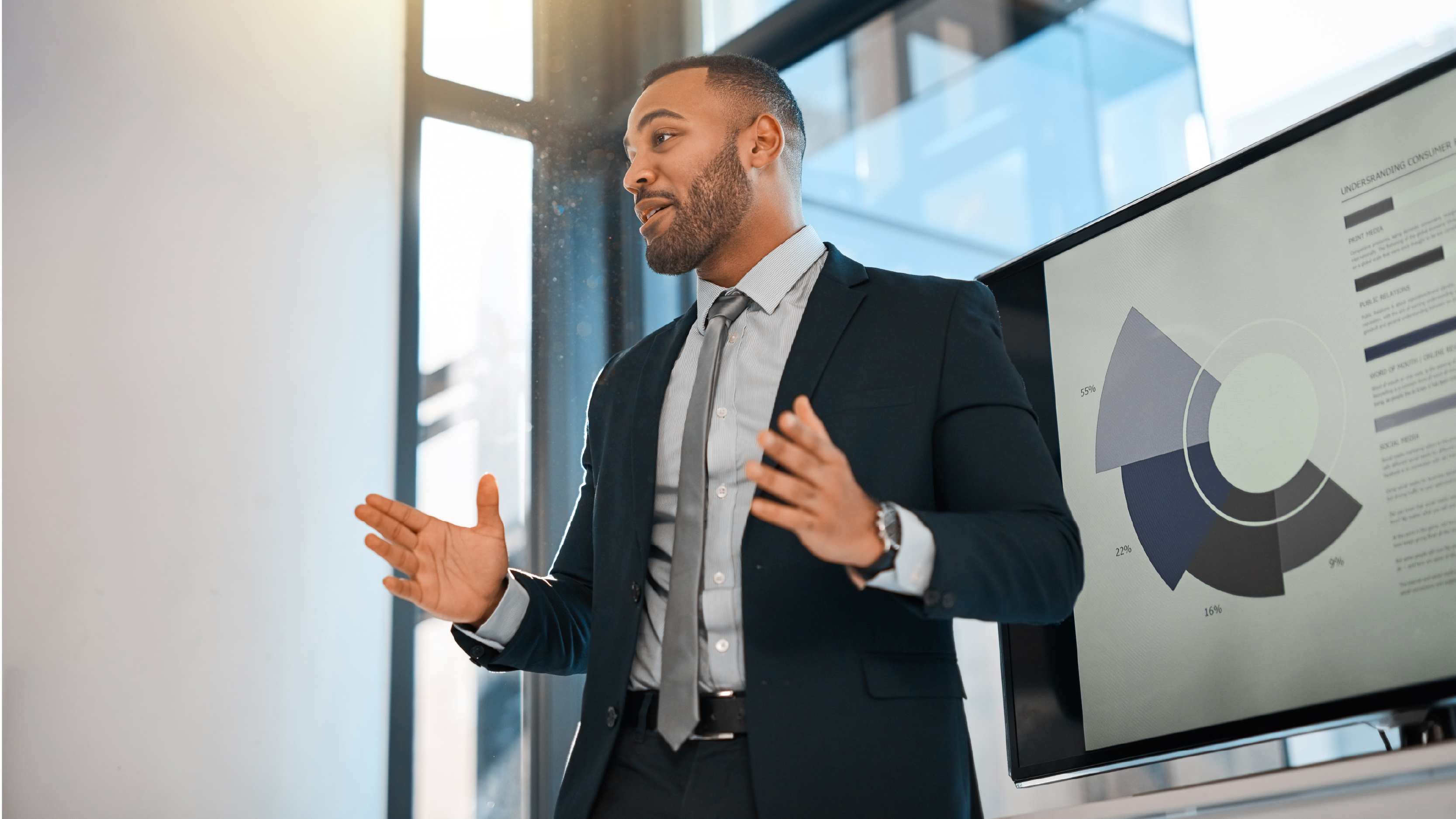 A business professional giving a presentation, standing beside a large screen displaying a segmented circular data chart in a modern office setting.