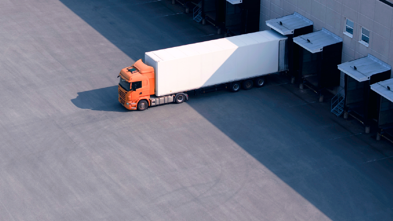 An orange truck parked in front of a large warehouse, showcasing its vibrant color against the industrial backdrop.