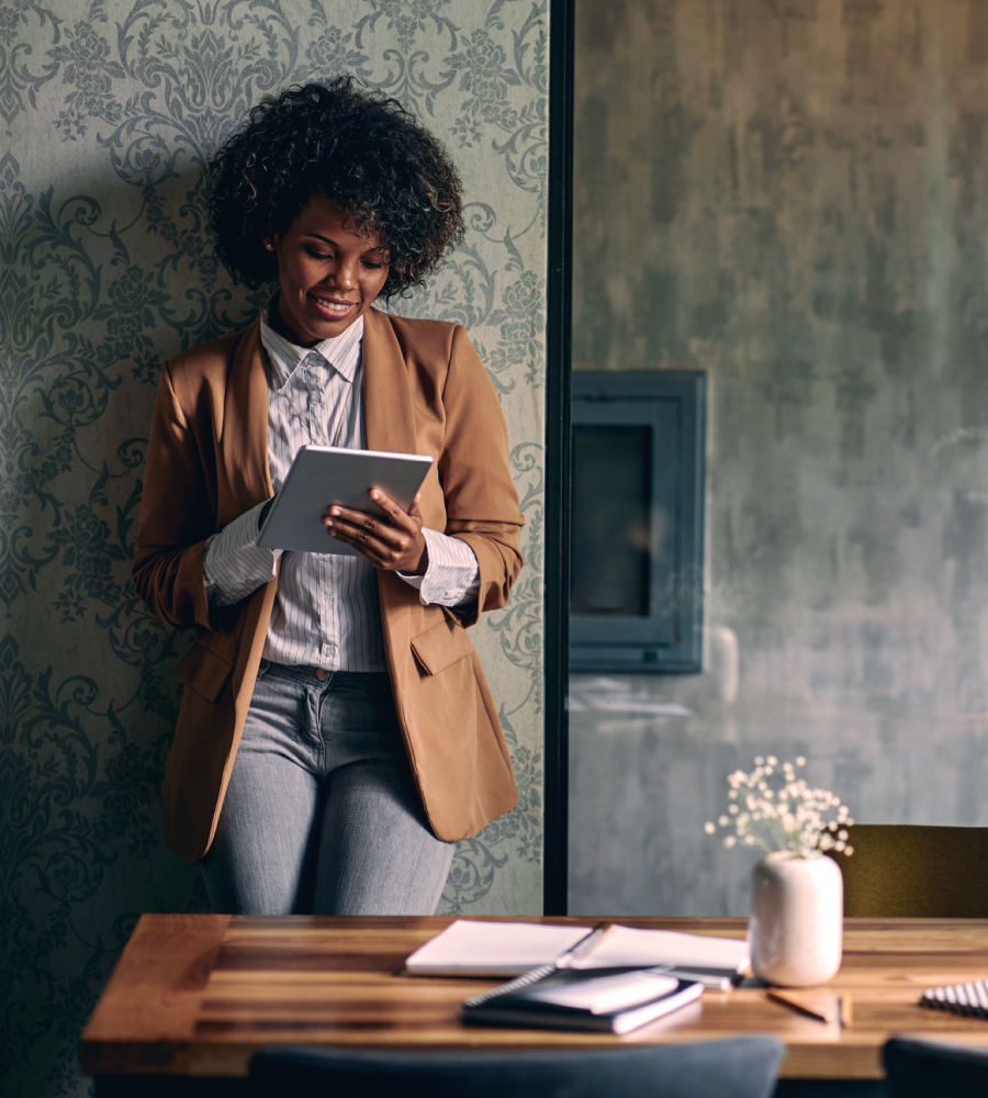 Smiling woman in a blazer leaning against a wall, using a tablet in a warmly lit room with a wooden table and notebooks in the foreground.