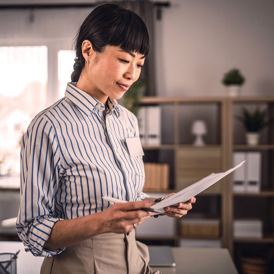 Professional woman in a striped shirt reviewing documents in a modern office setting, surrounded by shelves and natural light.