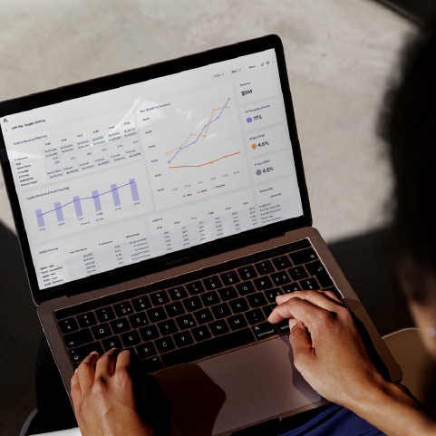 A person using a laptop displaying analytical charts and graphs on the screen, including line graphs, bar charts, and performance metrics, in a well-lit environment.