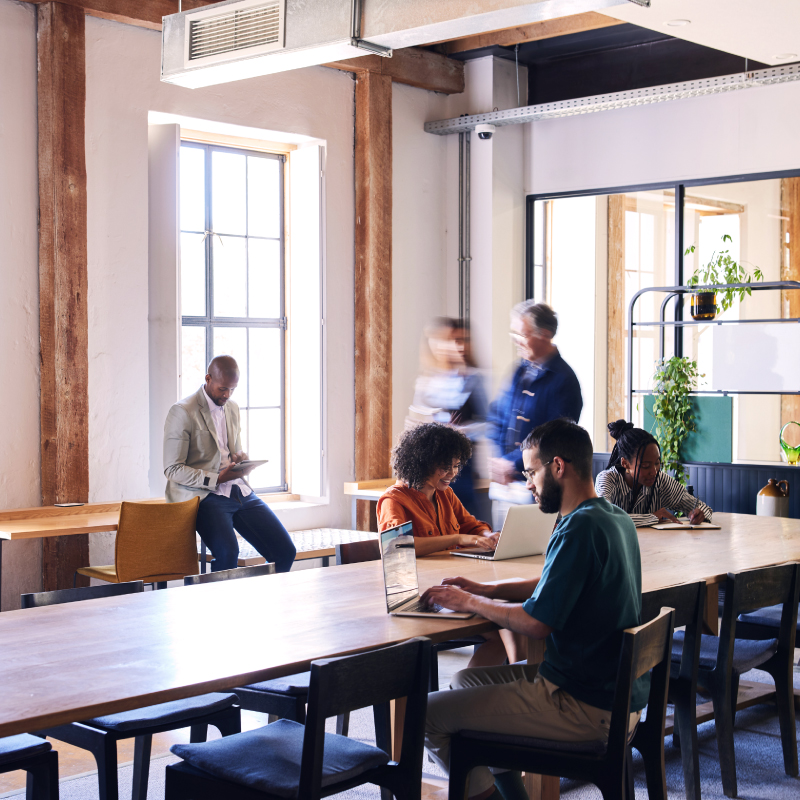 Modern coworking space with people working on laptops and tablets at a large wooden table, surrounded by natural light and greenery.