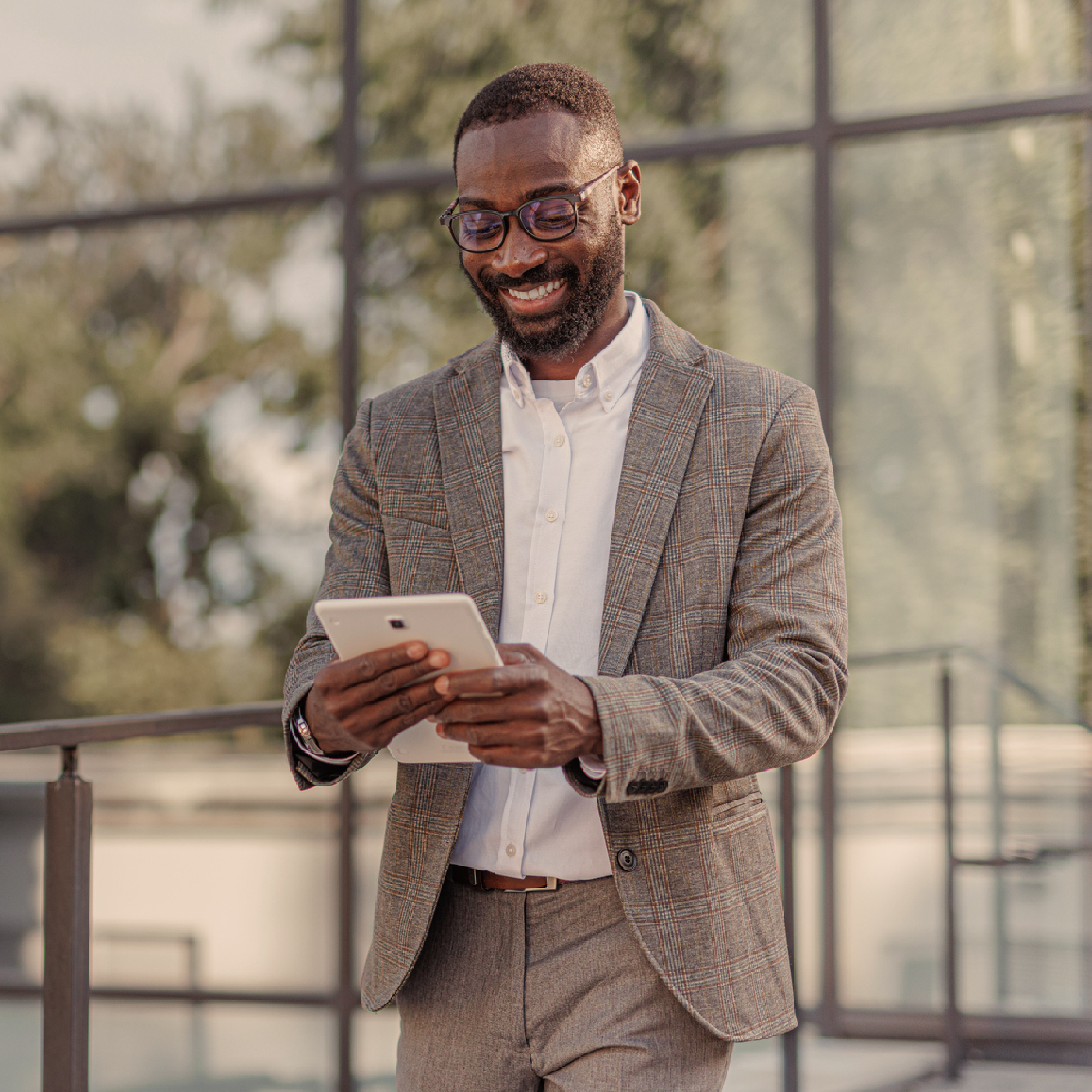 Smiling businessman in a plaid suit using a tablet outdoors, with a modern glass building in the background.