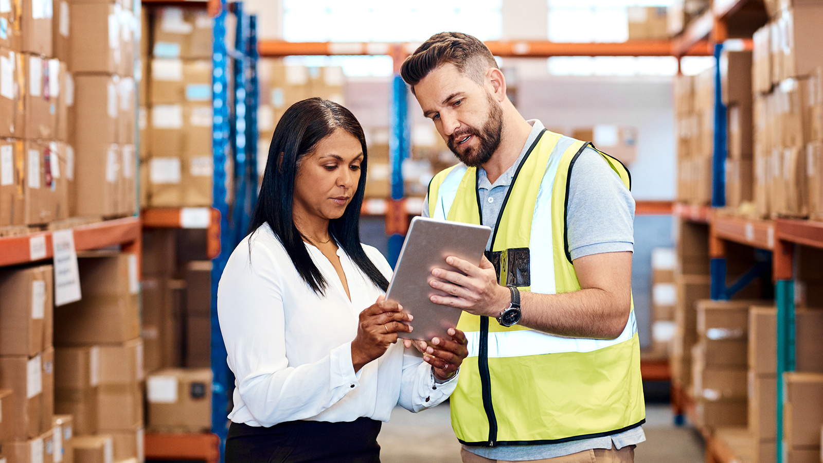 A warehouse environment where a man in a high-visibility vest is showing a tablet to a woman in a white blouse. Stacks of cardboard boxes line the shelves in the background.