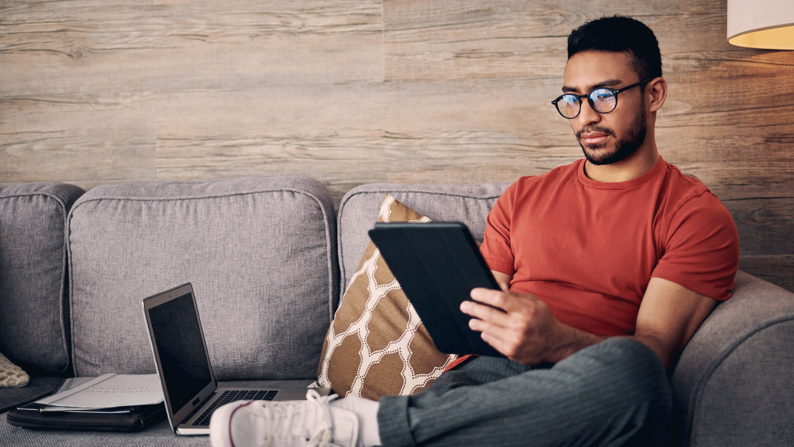 Man sitting on couch looking at a tablet with a laptop and paperwork next to him on the couch.