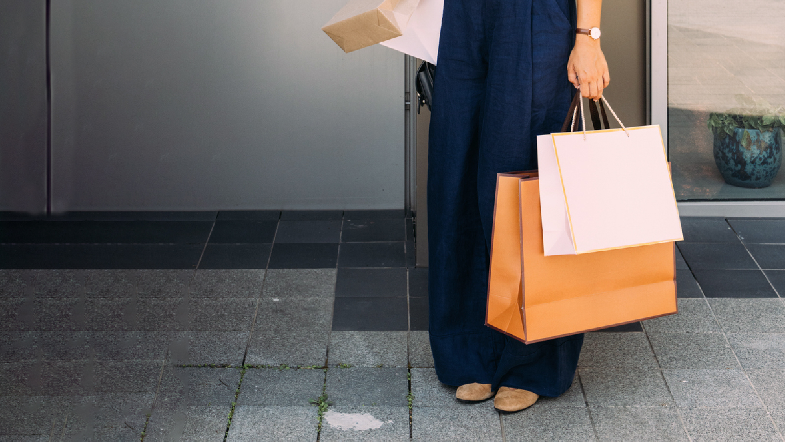 A woman in a blue jumpsuit stands confidently, holding several shopping bags in her hands.