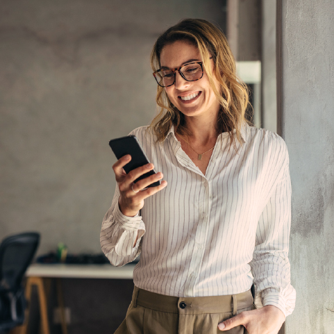 Smiling woman wearing glasses and a striped shirt, holding a smartphone while leaning against a wall in a casual office setting.