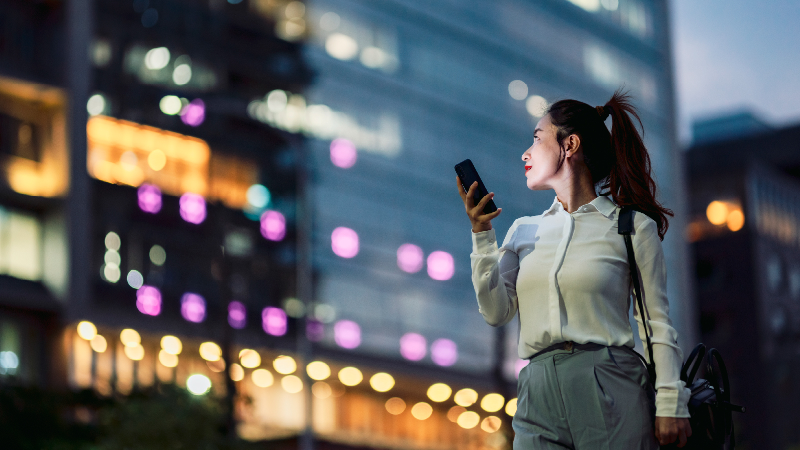 A woman is positioned in front of a building at night, using her phone while surrounded by the evening ambiance.