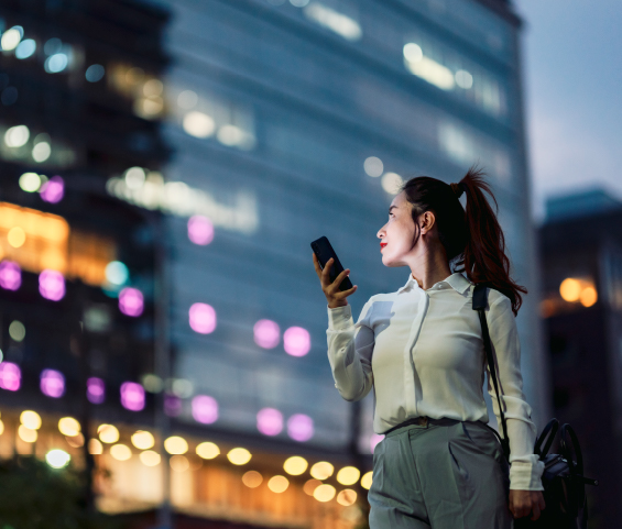 A woman is positioned in front of a building at night, using her phone while surrounded by the evening ambiance.