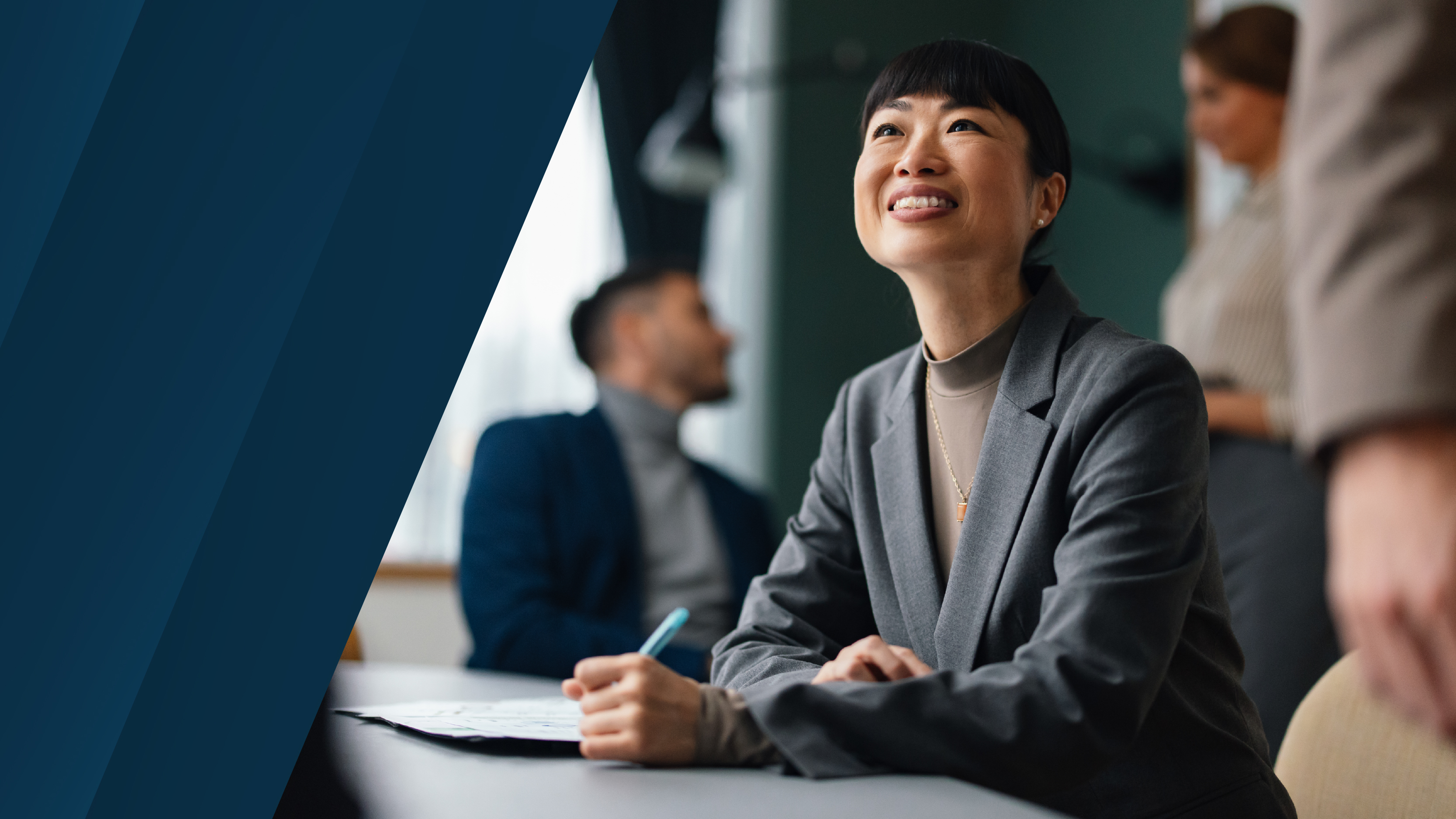 Image focused on a women at a table taking notes and smiling in a conference room full of people