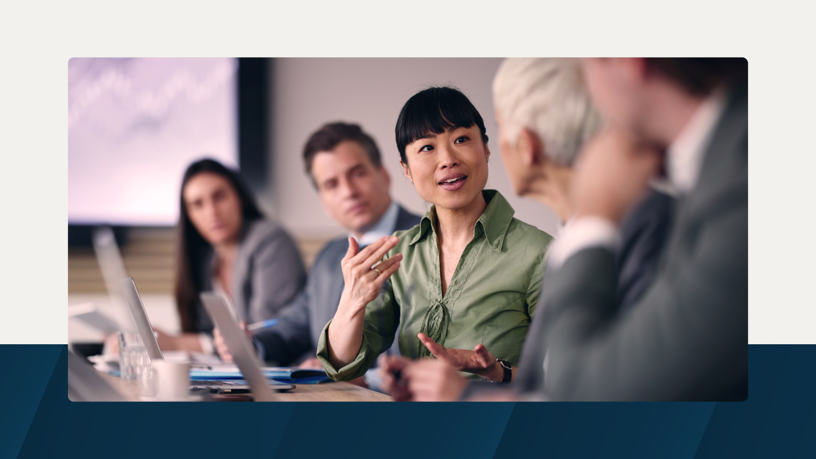 A woman in a green blouse leading a discussion in a professional meeting room with colleagues blurred in the background.