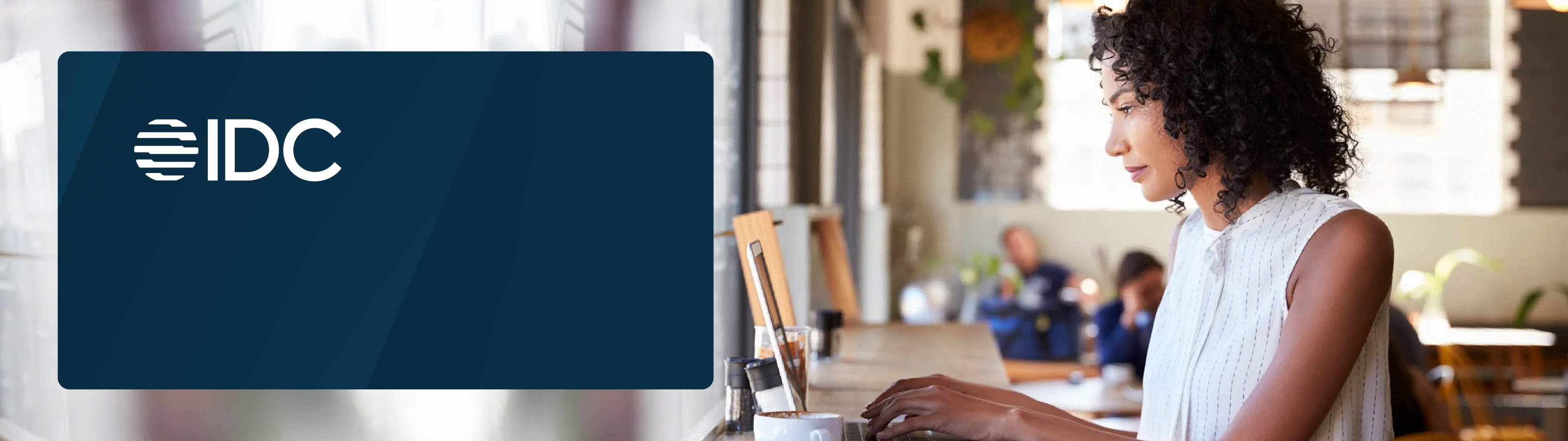 A woman working on a laptop at a café table, with an IDC logo overlay on the left side.